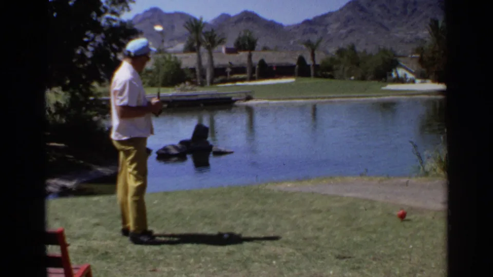 A 1968 Scottsdale golfer stands on the grass holding his club, with a red marker, pond, palms, and mountains behind him.