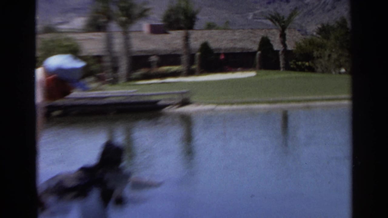 Blurred golfer near a pond with lawn, palms, and low buildings beyond.