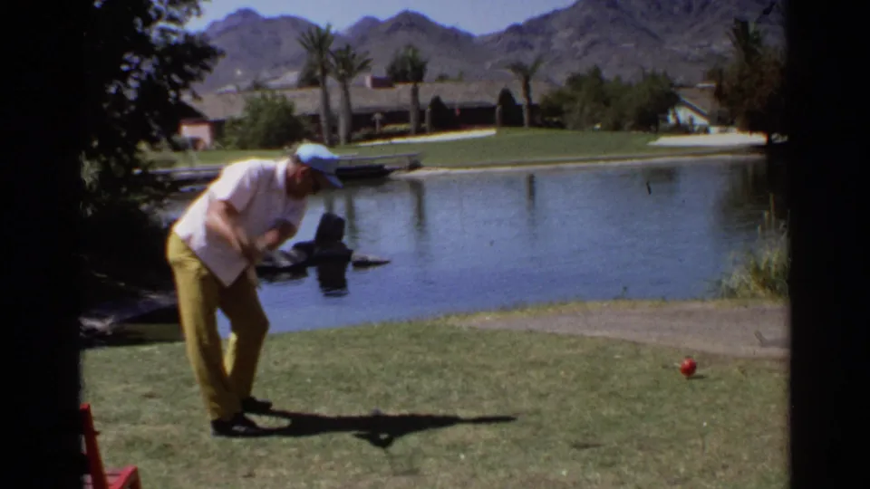 Adult golfer at a Scottsdale resort course, circa 1968, preparing to hit a red ball across a small pond with mountains behind.