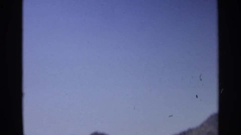 Brief 8mm sky frame from the golf sequence, showing blue sky, a sliver of mountain, dust marks, and dark gate edges.