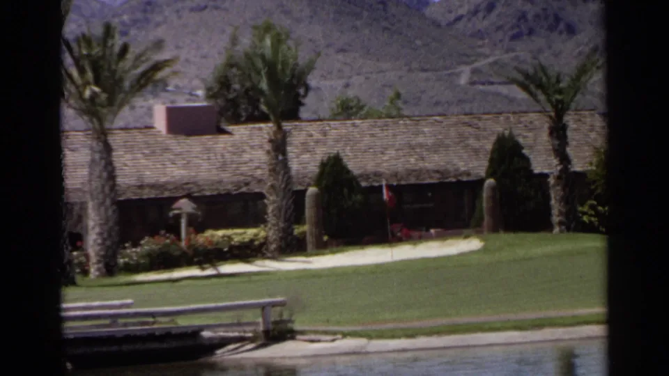 1968 Scottsdale view of a palm-lined golf course with a low shingled building, water feature, and desert mountains beyond.