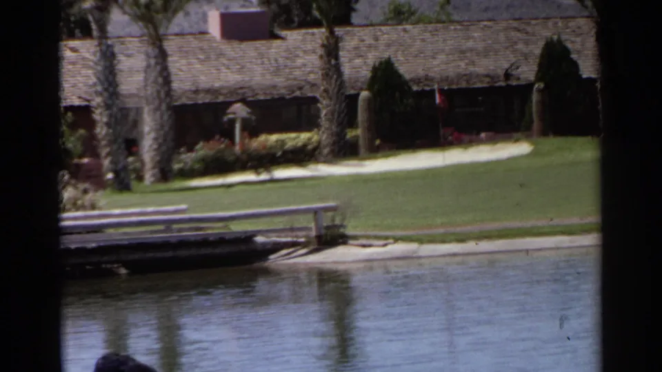 1968 Scottsdale golf-course view with bright grass, water in the foreground, palms, a walkway, and a low resort building.
