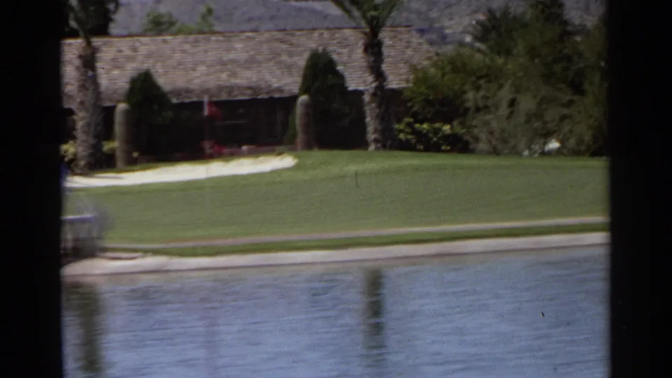 1968 Scottsdale frame showing a golf-course water edge, clipped lawn, palms, shrubs, and a low building in bright desert sun.