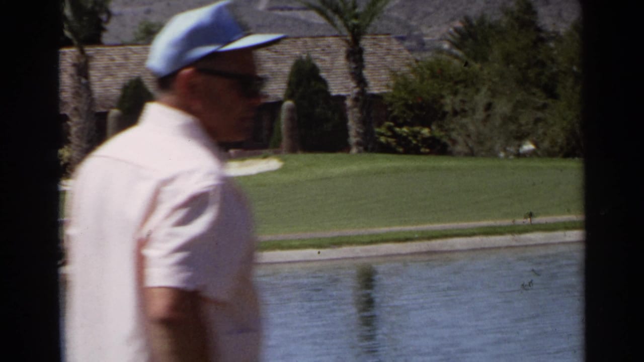 Older man in cap and sunglasses walks past a golf course water hazard.