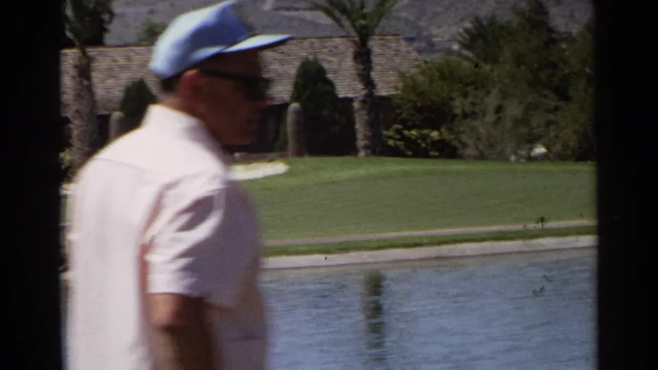 Older man in a light blue cap and sunglasses walks beside a sunny Scottsdale golf course water hazard in 1968 home-movie footage.