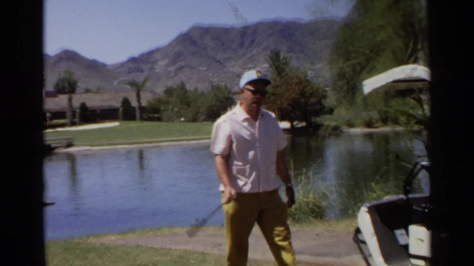 Man in cap and sunglasses stands near a golf cart with a club beside a water hazard on a Scottsdale course.