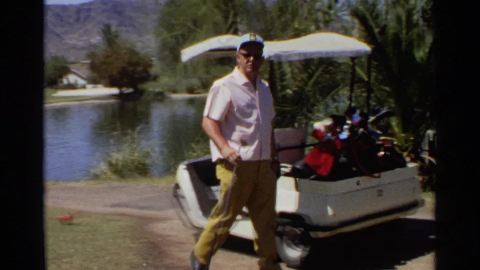 Older golfer stands beside a cart carrying clubs and bags near a sunny Scottsdale water hazard, with palms and mountains visible.