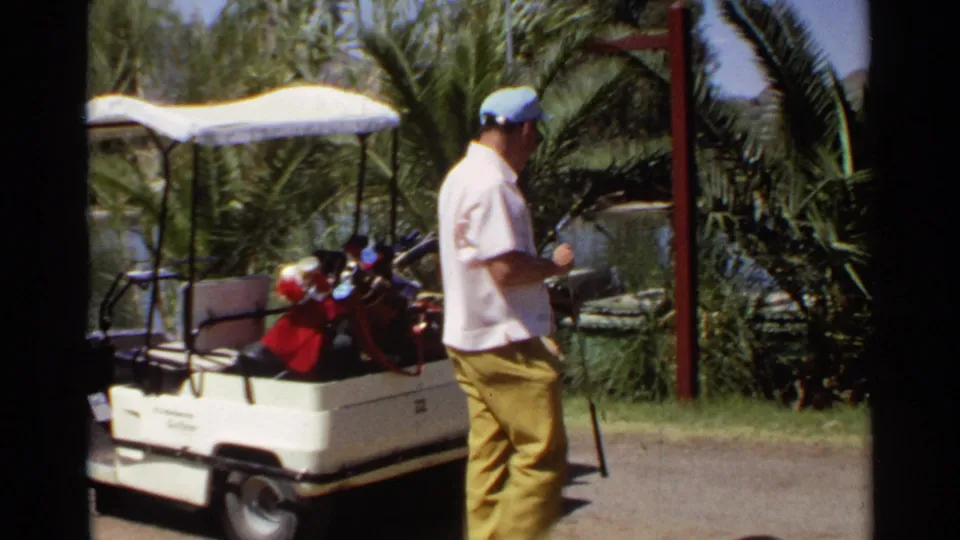Man in mustard trousers carries a golf club away from a cart amid palms and course landscaping in 1968 Scottsdale footage.