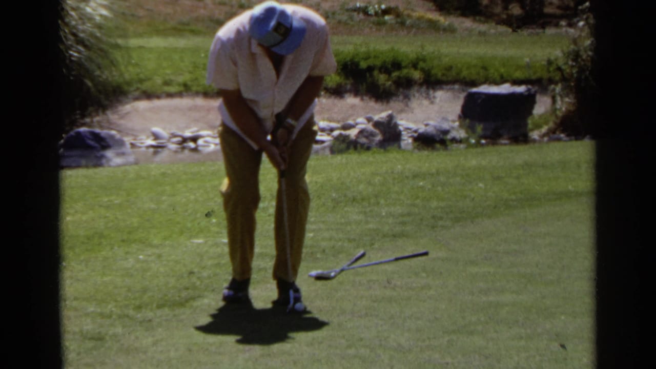 Man bends over a putter on a sunny golf green with another club on the grass.