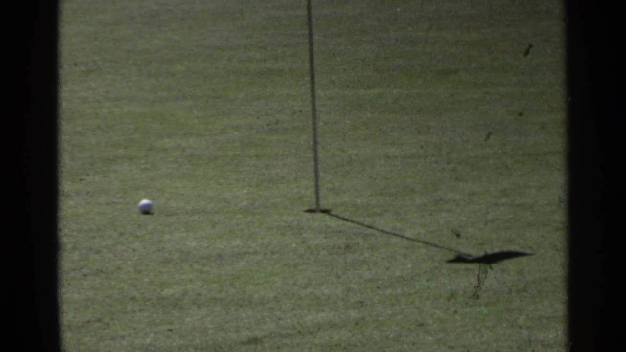 The ball approaches the cup and flagstick on the Scottsdale golf green, with the flag shadow visible on the grass.