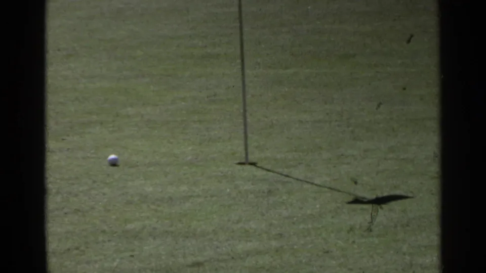 The ball approaches the cup and flagstick on the Scottsdale golf green, with the flag shadow visible on the grass.