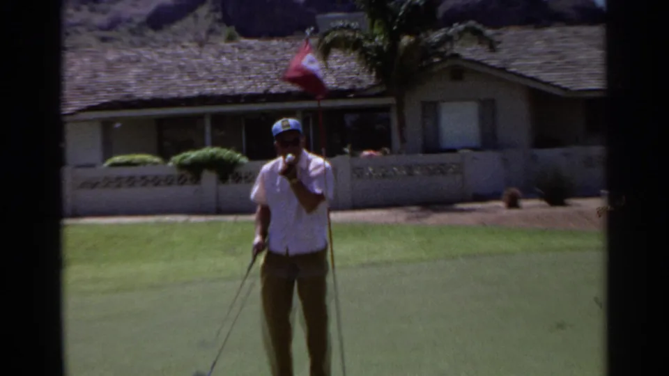 An adult golfer in a white shirt and blue cap stands near the putting green in Scottsdale, with a red flag and low home behind him.