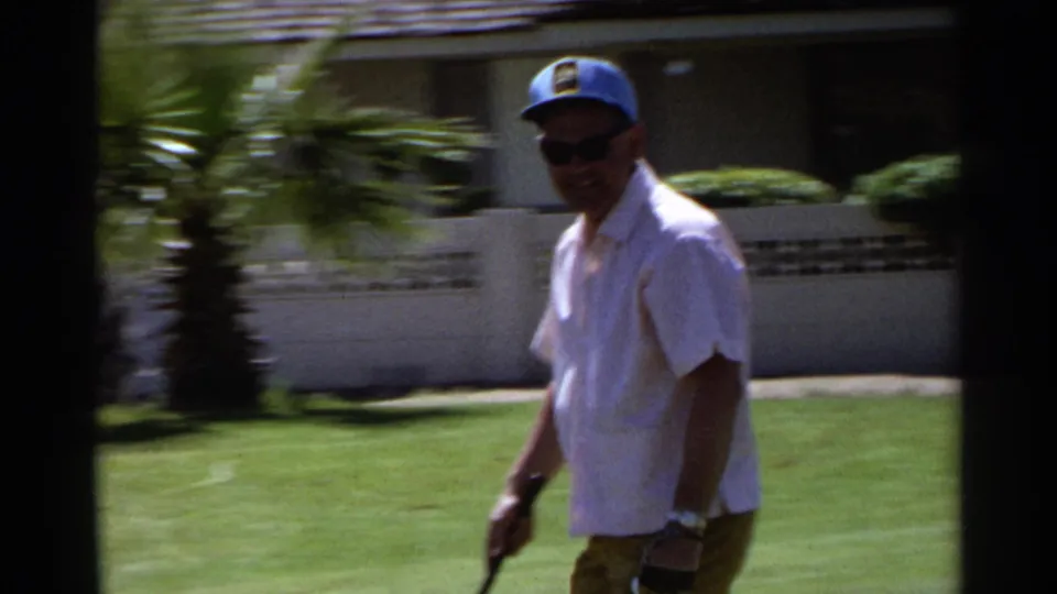 A man in a white short-sleeve shirt, khaki pants, sunglasses, and blue cap walks across a Scottsdale golf green with a putter.