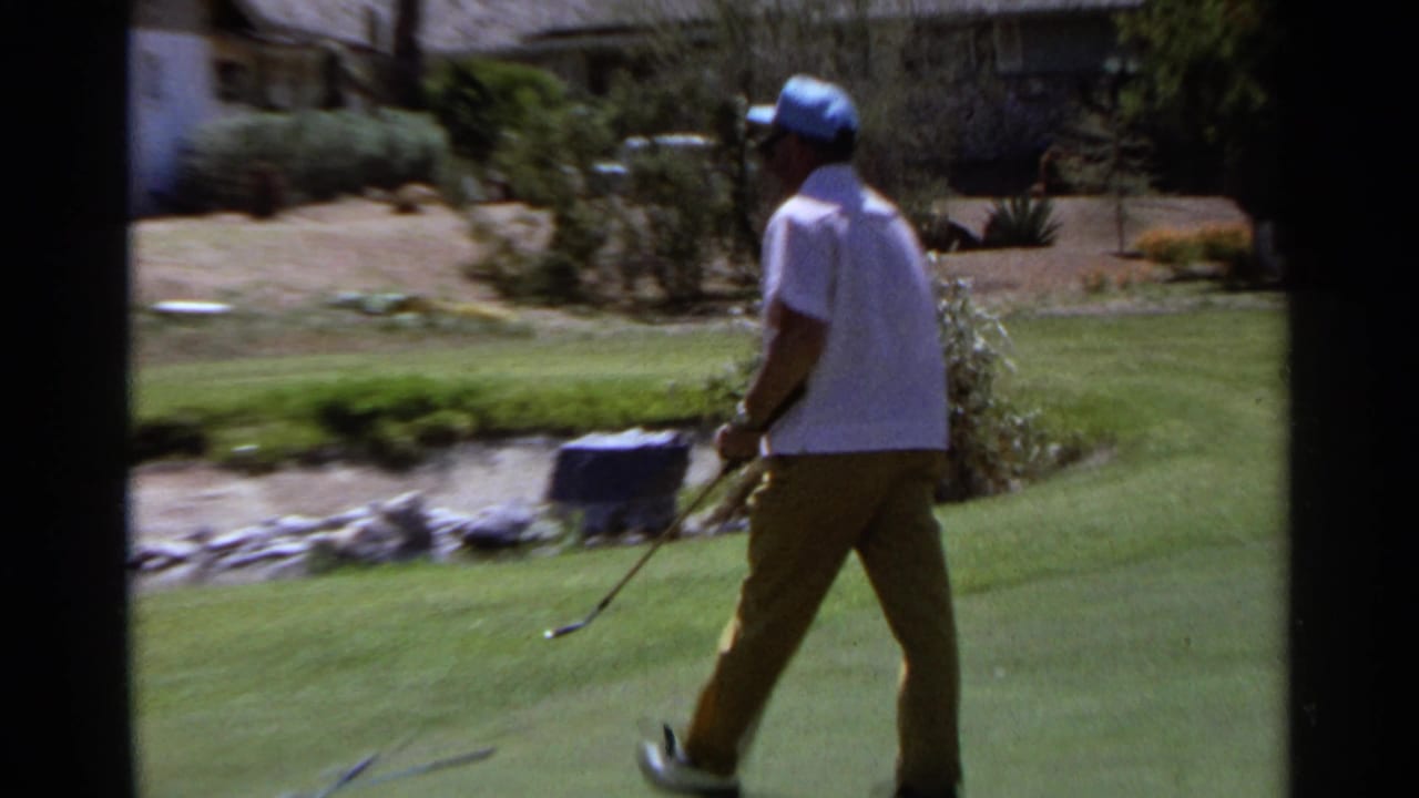 Golfer in a white shirt and blue cap walking on a green with a golf club.