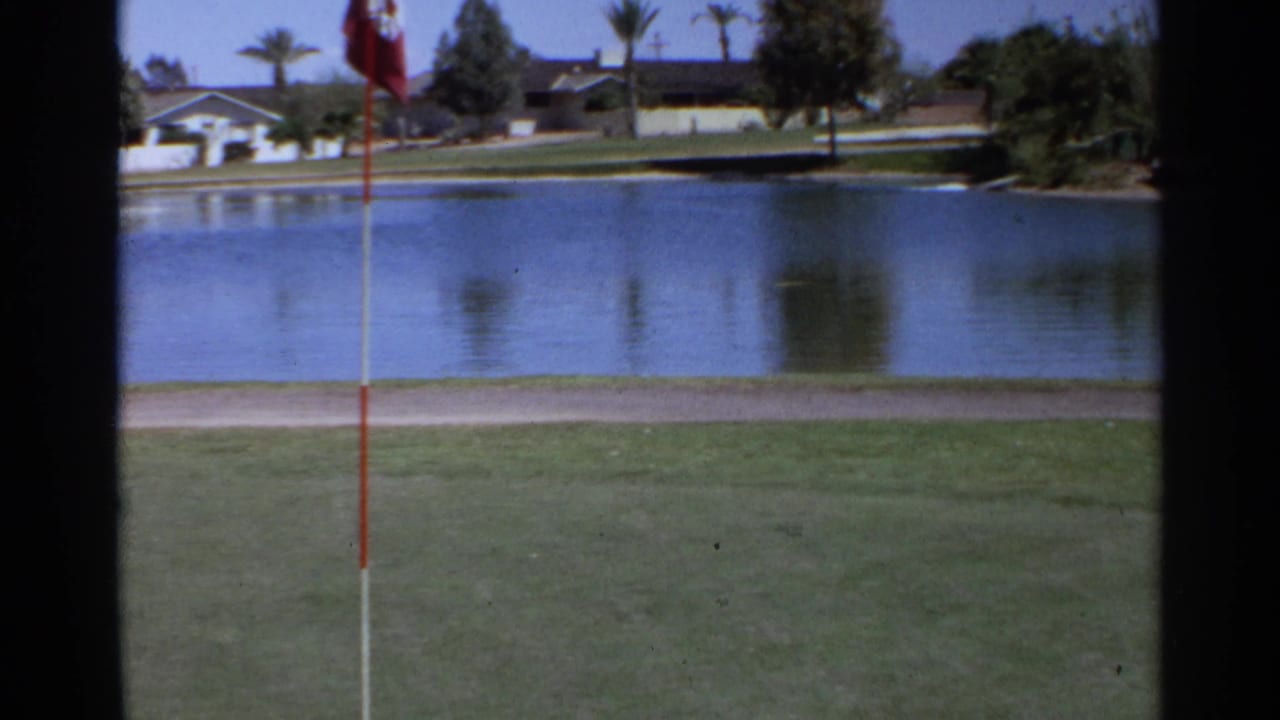 Golf flag beside a water hazard with grass and houses beyond.
