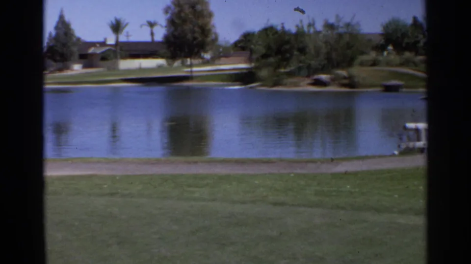 1968 Scottsdale golf course frame looking across a pond toward palms, trees, fairway grass, and distant low houses.