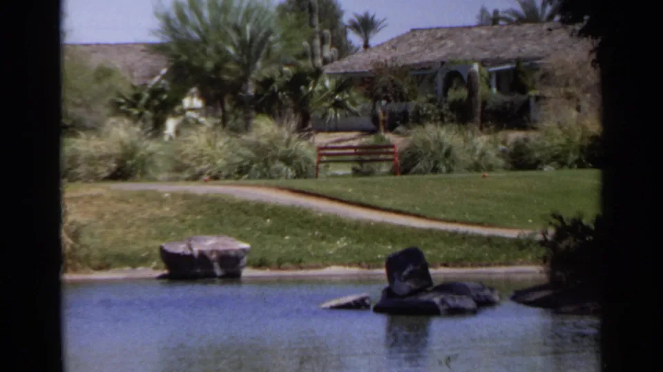 1968 Scottsdale pond view showing rocks in the water, a curved path, palms, a bench, lawn, and a low building.