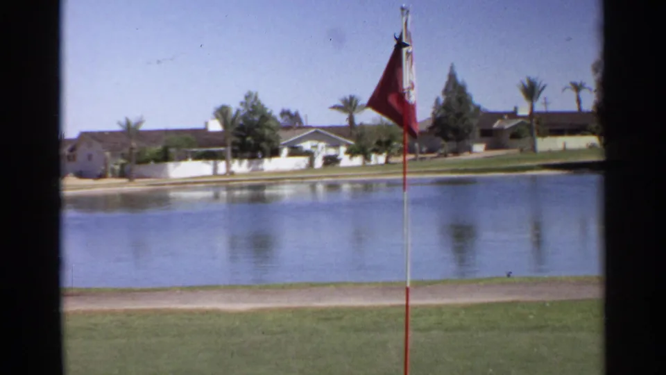1968 Scottsdale golf-course view with a red flag in the foreground, pond water, grass, path, palms, and low buildings beyond.