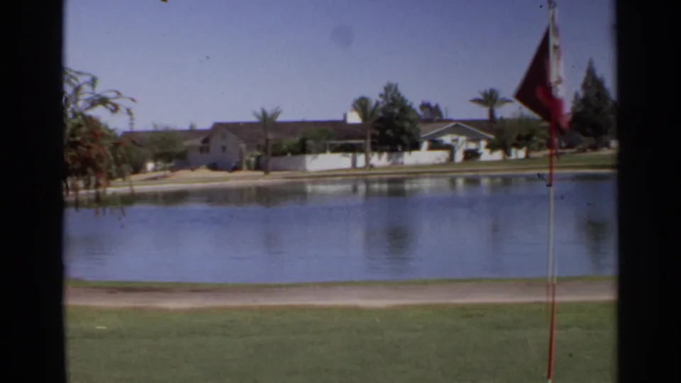 1968 Scottsdale golf-course pond with a red flag at right, smooth water, lawn, palms, and low buildings across the pond.