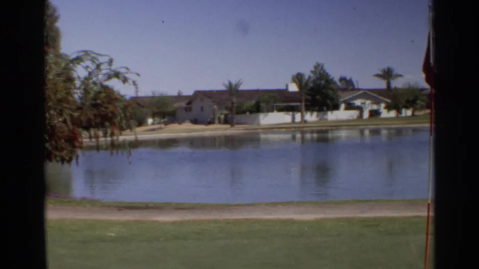 1968 Scottsdale home-movie frame showing a calm pond, grass, a dirt path, distant residential houses, palm trees, and water reflections.