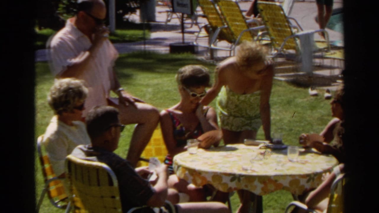 Adults and children sit around a floral poolside table while a man stands nearby.