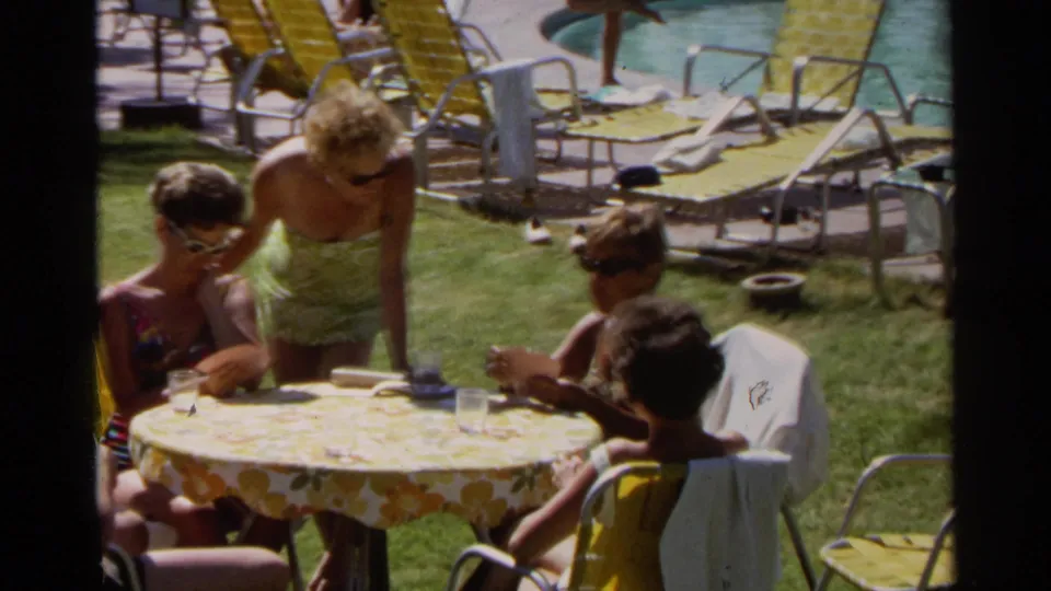 Adults and children remain around the floral table at a Scottsdale resort pool, with 1960s lounge chairs and pool activity behind them.