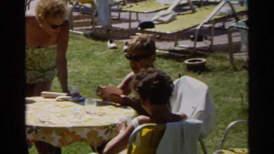 A woman in sunglasses stands by children at a floral poolside table in the Scottsdale home-movie resort scene.