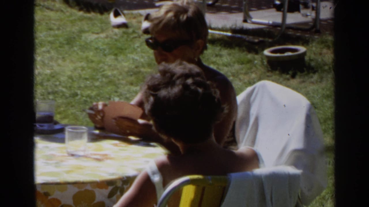Adults in sunglasses sit around a lawn table with drinks and a floral tablecloth.