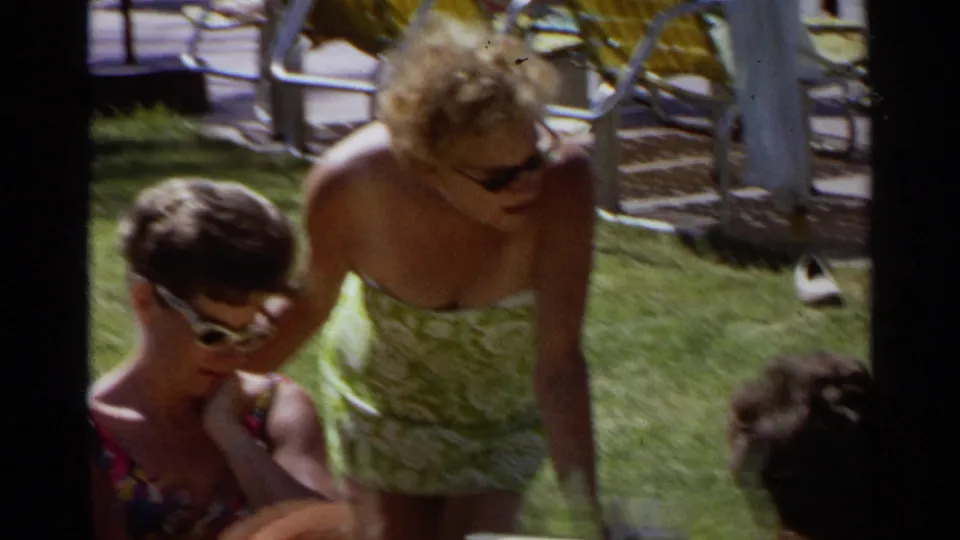 1968 Scottsdale frame with a woman in a green patterned swimsuit standing beside seated adults in sunglasses on a lawn.