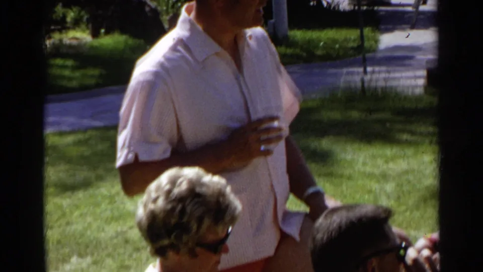 1968 Scottsdale frame with a man in a light short-sleeve shirt holding a drink near seated adults on a sunlit lawn.