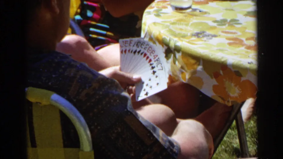 A seated adult holds playing cards at a yellow floral table during a sunny 1968 Scottsdale backyard card game.