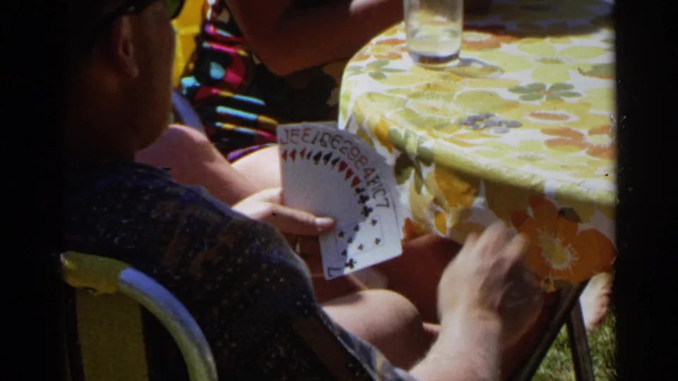 Players sit around a bright yellow floral table as one adult holds cards in 1968 Scottsdale backyard footage.