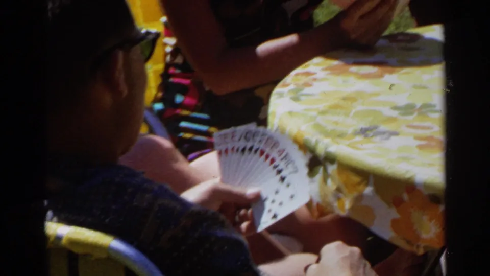 An adult card player sits at a yellow floral table with another seated person nearby in sunny Scottsdale home-movie footage from 1968.