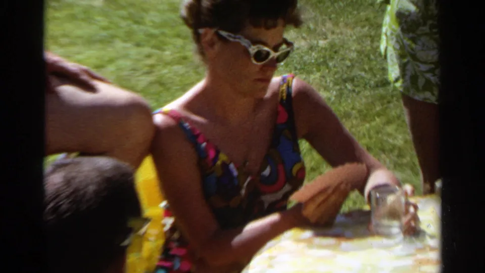 Scottsdale 1968 home-movie still of a woman in sunglasses looking down at cards at a sunny lawn table.