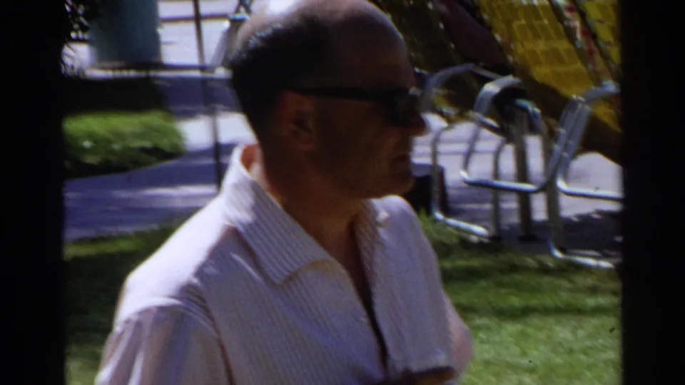 1968 Scottsdale frame of an adult man in sunglasses and a light striped shirt holding a drink near patio chairs on a sunny lawn.