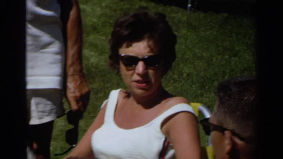 1968 Scottsdale home-movie frame showing a seated woman in sunglasses and a white tank top talking with adults on a sunny lawn.