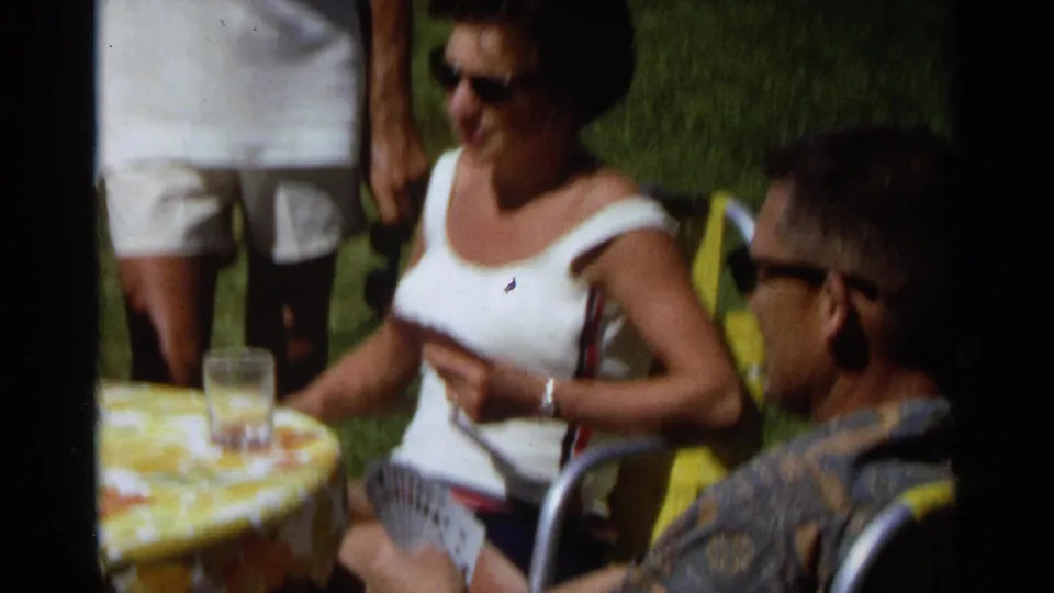 1968 Scottsdale frame of adults seated on lawn chairs around a yellow floral tablecloth during an outdoor card game.