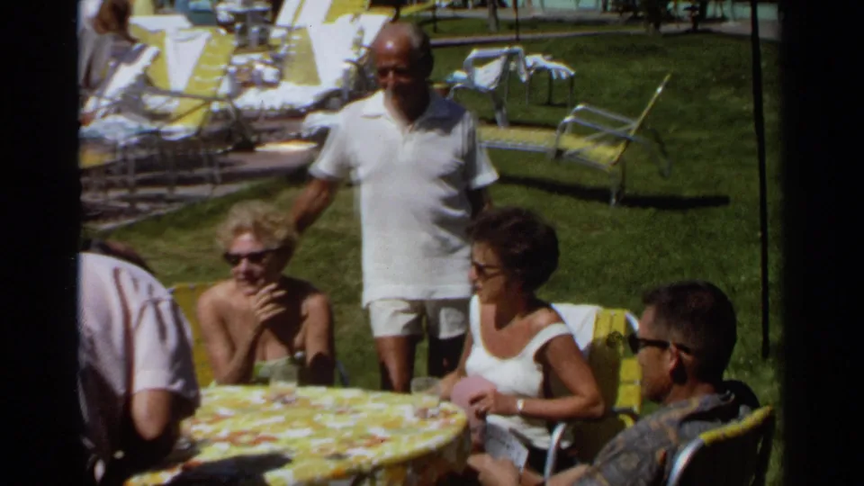 1968 Scottsdale home-movie frame with a man in a white shirt standing near adults seated around an outdoor card table.