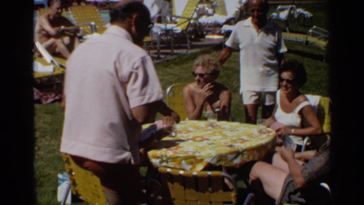 Adults gather around a yellow floral table outdoors while a man stands serving drinks.