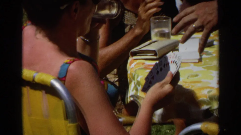 In the final frame, a woman holds playing cards during an outdoor table game with glasses and a deck on the floral tablecloth.