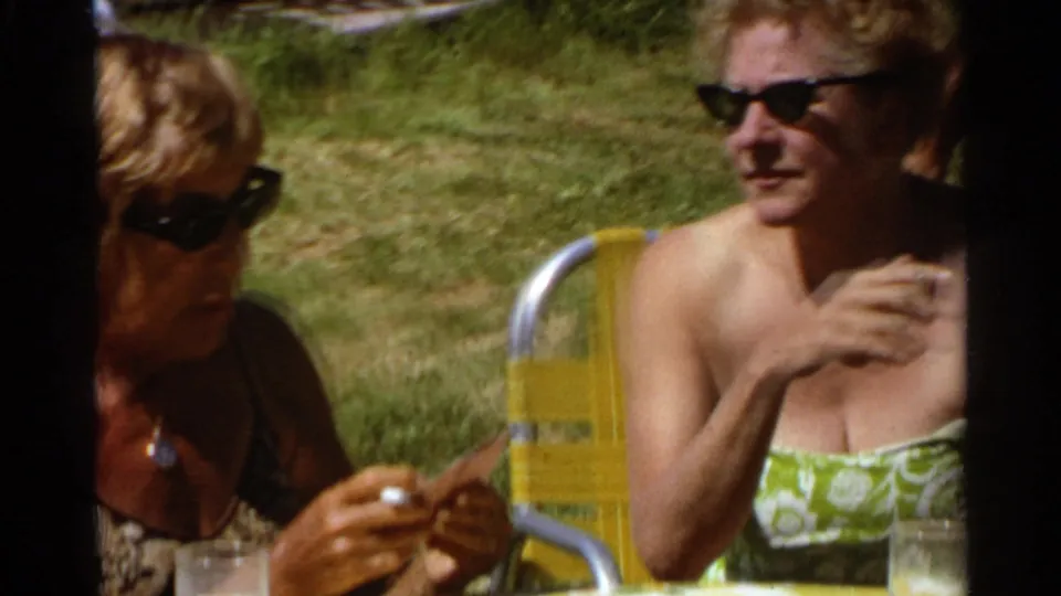 1968 Scottsdale frame showing two women in sunglasses seated on a grassy lawn beside a yellow chair and outdoor table.