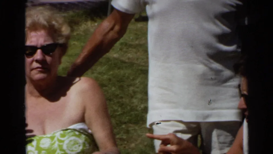 1968 Scottsdale home-movie frame with a seated woman in sunglasses and a standing adult partly blocking the grassy background.