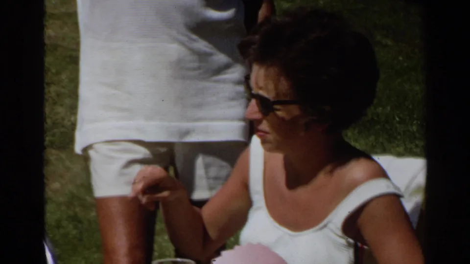 1968 Scottsdale frame of a woman in sunglasses and a white swimsuit seated on a lawn with a standing adult partly in front.