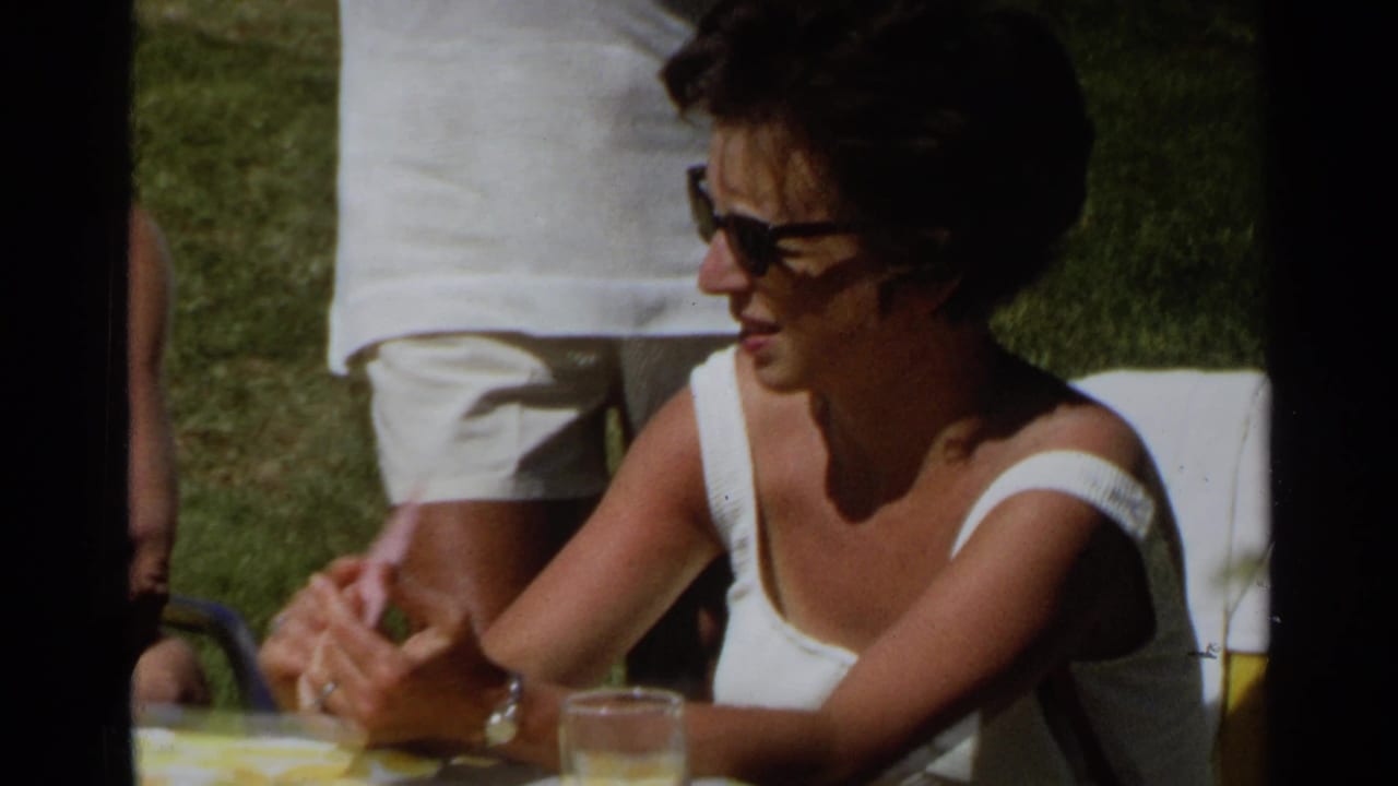 A woman in dark sunglasses sits at an outdoor table in Scottsdale, Arizona, with a glass and standing figures nearby.