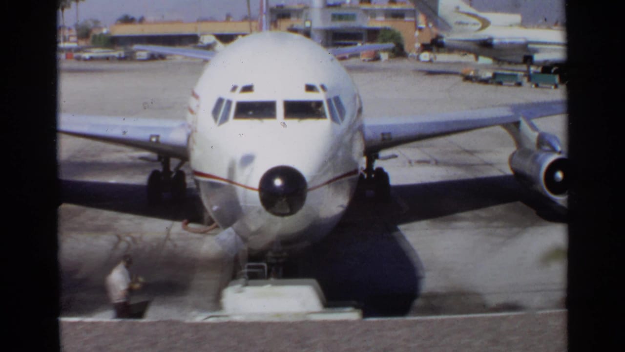 The camera frames the nose and cockpit windows of a white commercial airplane parked on a tarmac in the 1968 travel sequence.