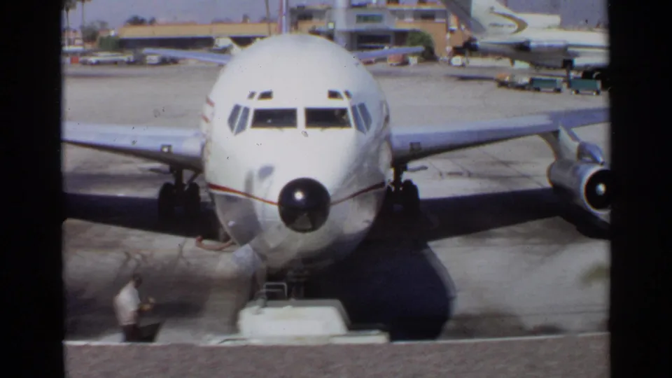The camera frames the nose and cockpit windows of a white commercial airplane parked on a tarmac in the 1968 travel sequence.