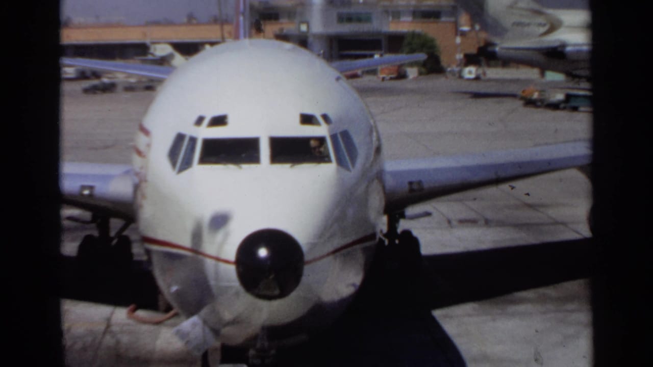 A closer home-movie view shows the front of a white commercial airplane, with cockpit windows, nose cone, wing, and tarmac visible.
