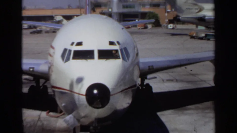 A closer home-movie view shows the front of a white commercial airplane, with cockpit windows, nose cone, wing, and tarmac visible.