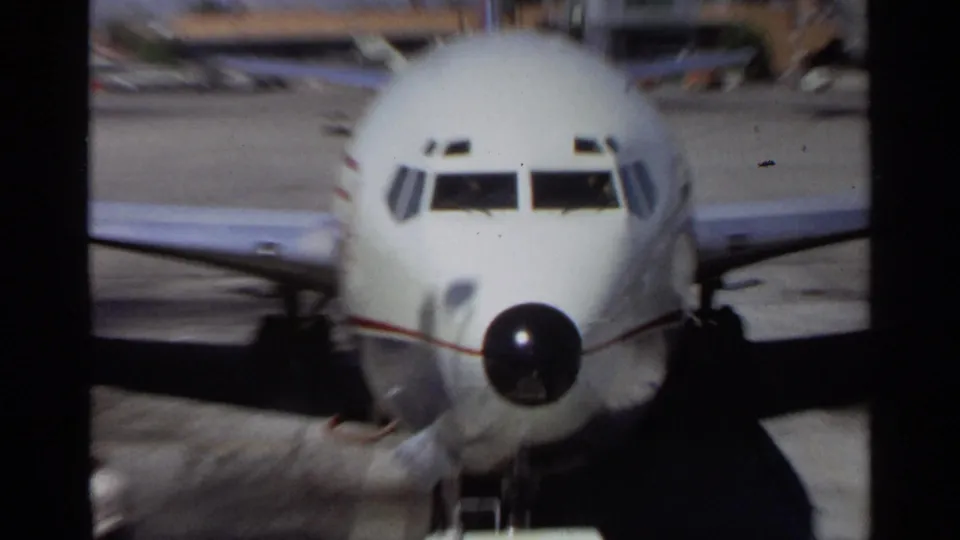 Wider 1968 Scottsdale home-movie view of the white airplane facing camera, with wings, black nose cone, tarmac, and airport buildings behind.