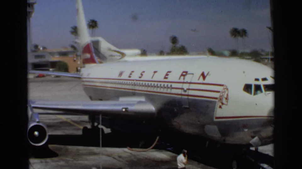 Parked Western Airlines jet seen from the side in 1968 Scottsdale footage, with wing, engine, tarmac, and worker visible.